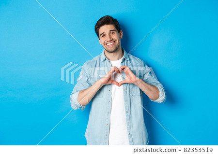 Handsome man smiling, showing heart gesture and looking at camera, saying I love you, standing against blue background 82353519