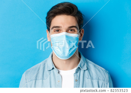Concept of covid-19, pandemic and quarantine. Close-up of happy guy in medical mask looking at camera, standing over blue background Concept of covid-19, pandemic and quarantine. Close-up of happy guy in medical mask looking at camera, standing over blue background 82353523