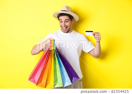 Concept of vacation and finance. Happy man shopper looking at shopping bags satisfied, showing credit card, standing against yellow background Concept of vacation and finance. Happy man shopper looking at shopping bags satisfied, showing credit card, standing against yellow background 82354425