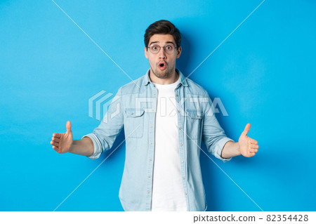 Surprised handsome man holding something big, showing large size with amazed face, standing over blue background 82354428