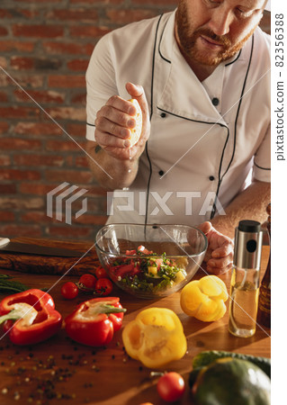 Young Caucasian red-bearded man, chef cooking fresh vegetable salad in cafe, restaurant kitchen. Concept of a correct, healthy diet. 82356388