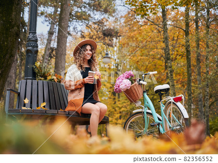 Stylish smiling woman with curly hair in stylish beige hat and cardigan sitting on bench with tasty coffee or tea in beautiful autumn park. Concept of riding bicycles in city park. Stylish smiling woman with curly hair in stylish beige hat and cardigan sitting on bench with tasty coffee or tea in beautiful autumn park. Concept of riding bicycles in city park. 82356552