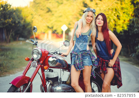 Young beautiful women ride a retro motorcycle. A talented girl in a blue overalls. They are standing near a motorcycle, funny go to the road. sale of spare parts. Place for text 82357855