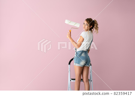 Beautiful woman is standing on the ladder and is painting the wall to pink color with a roller on her hand. Beautiful woman is standing on the ladder and is painting the wall to pink color with a roller on her hand. 82359417
