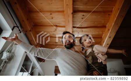 Low angle view of mature father with small daughter standing indoors at home, holding and hugging. Low angle view of mature father with small daughter standing indoors at home, holding and hugging. 82359892