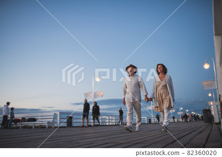 Low angle view of happy senior couple walking outdoors on pier by sea at dusk, holding hands. 82360020