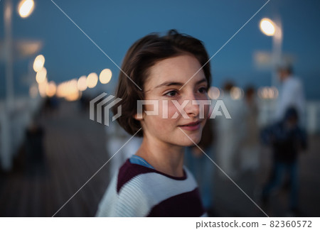 Portrait of preteen girl looking aside outdoors on pier by sea at dusk, holiday concept. 82360572