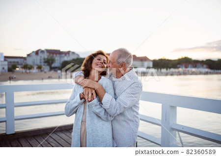 Happy senior couple hugging outdoors on pier by sea, looking at each other. 82360589