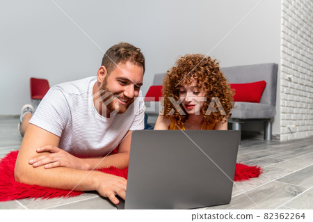 A Young couple lovers or friends laying on a red pluffy carpet and using laptop. 82362264