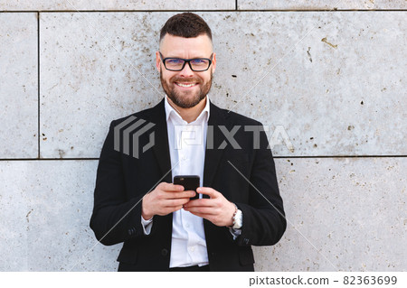 Positive urban business professional man in stylish suit using smartphone while standing outside Positive urban business professional man in stylish suit using smartphone while standing outside 82363699
