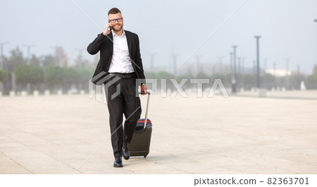 Young confident businessman in formal black suit pulling suitcase luggage after landing in airport Young confident businessman in formal black suit pulling suitcase luggage after landing in airport 82363701