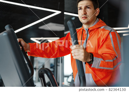 Portrait of a young man in orange windbreaker workout on a fitness machine at gym. 82364130
