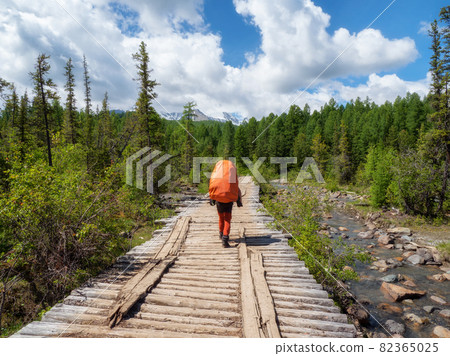 A male tourist with a large orange backpack walks along an old wooden bridge against the background of a coniferous forest and mountains in the distance A male tourist with a large orange backpack walks along an old wooden bridge against the background of a coniferous forest and mountains in the distance 82365025