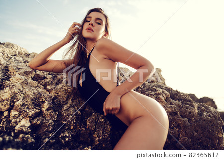 Young beautiful sexy female model in black bikini standing in sea near huge rock 82365612