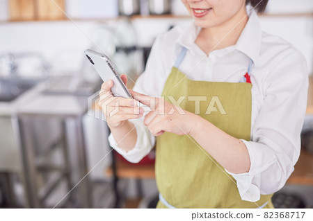 A young woman in an apron operating a smartphone in the kitchen A young woman in an apron operating a smartphone in the kitchen 82368717