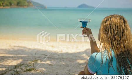 The woman is holding a glass of blue curacao cocktail, on beach azure blue sea of a tropical island. Blurred beautiful seaside background. The woman is holding a glass of blue curacao cocktail, on beach azure blue sea of a tropical island. Blurred beautiful seaside background. 82376167