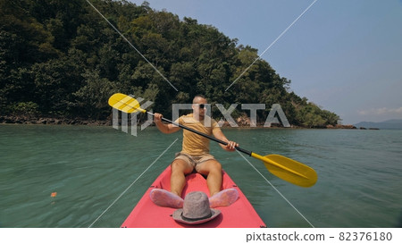 Man with sunglasses and hat rows pink plastic canoe along sea against green hilly islands with wild jungles. Traveling to tropical countries. 82376180