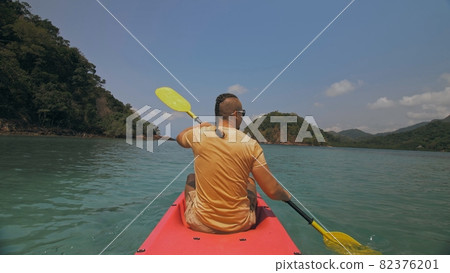 Man with sunglasses and hat rows pink plastic canoe along sea against green hilly islands with wild jungles. Traveling to tropical countries. Man with sunglasses and hat rows pink plastic canoe along sea against green hilly islands with wild jungles. Traveling to tropical countries. 82376201