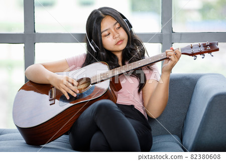 Portrait shot of young teenager enjoying playing the acoustic guitar and wearing a headset. Professional junior guitarist sitting on the couch and holding an instrument with windows background 82380608