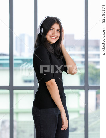 Portrait shot of cute smiling young Thai-Turkish teenager enjoying listening to music with a headset while looking at camera. Junior girl with casual black clothes holding a smartphone to play music 82380873