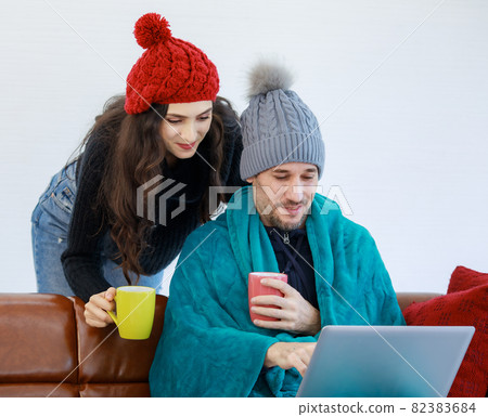 Caucasian male and female lover married couple in wool sweater jacket and knitted hat cover by warm blanket drink hot beverage surfing browsing internet shopping online together at home in winter 82383684