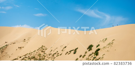 A wide panorama of a large dune with a small silhouette of people walking on it against the blue sky A wide panorama of a large dune with a small silhouette of people walking on it against the blue sky 82383736