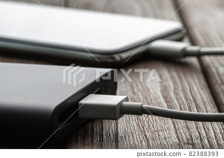 The smartphone is charging from external powerbank with cable on a brown wooden background. Closeup, selective focus The smartphone is charging from external powerbank with cable on a brown wooden background. Closeup, selective focus 82388393
