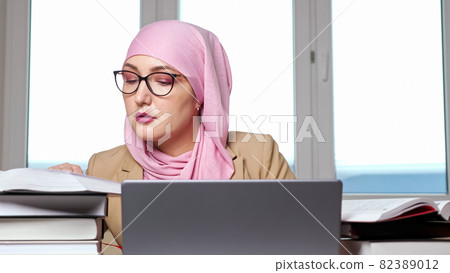 Woman in hijab and glasses typing on laptop looking into books Woman in hijab and glasses typing on laptop looking into books 82389012