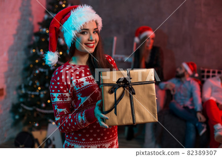 Portrait of a young woman with a gold giftbox at home on the foreground. Beautiful brunette in a Santa hat, red costume with deers Portrait of a young woman with a gold giftbox at home on the foreground. Beautiful brunette in a Santa hat, red costume with deers 82389307