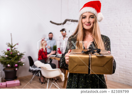 Portrait of a young woman with a gold giftbox at home on the foreground. Beautiful blonde in a Santa hat and black dress 82389799