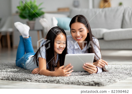Excited asian mother helping her daughter with lesson on digital tablet, lying together on floor carpet 82398462