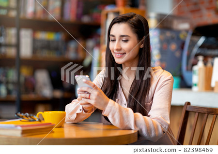 Young happy arab woman using mobile phone, sitting in city cafe and surfing social networks, drinking coffee and resting 82398469
