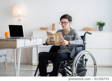 Focused impaired teen boy in wheelchair reading textbook, studying from home during covid quarantine Focused impaired teen boy in wheelchair reading textbook, studying from home during covid quarantine 82398741