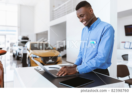 Positive black car salesman using laptop computer at work desk in auto dealership, copy space 82398760
