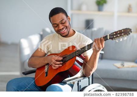 Positive paraplegic black man in wheelchair playing guitar, enjoying music at home Positive paraplegic black man in wheelchair playing guitar, enjoying music at home 82398788