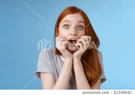 Shocked speechless gasping young redhead girl staring impressed stunned watching important moment tv series biting fingers open mouth shook standing excited blue background anticipating Shocked speechless gasping young redhead girl staring impressed stunned watching important moment tv series biting fingers open mouth shook standing excited blue background anticipating 82399692