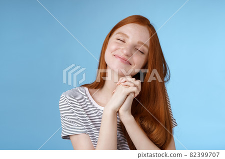 Charming delighted happy dreamy redhead girlfriend ginger long hair tilting head close eyes smiling pleased daydreaming thinking about sweet tender memories press palms together, blue background 82399707
