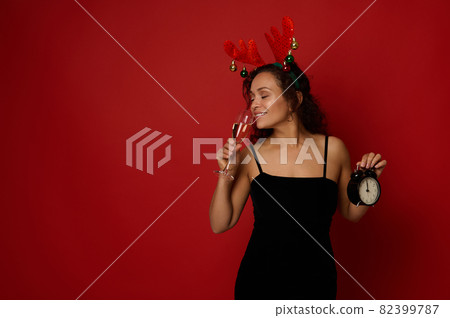 Beautiful Hispanic woman wearing a deer antler hoop and black velvet dress holding an alarm clock and drinking sparkling wine from a flute with champagne, isolated on red background. Christmas concept 82399787