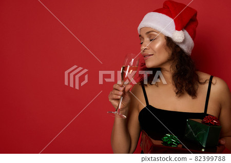 Isolated close-up portrait on red background with copy space of attractive woman in with evening shining make-up, wearing Santa hat, drinking sparkling wine from flute and holding Christmas gift boxes 82399788