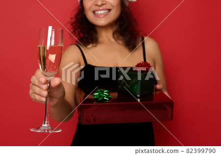 Soft focus on champagne flute with sparkling wine on the background of blurred cheerful smiling woman with beautiful toothy smile, holding Christmas gift boxes wrapped in glitter red green gift paper Soft focus on champagne flute with sparkling wine on the background of blurred cheerful smiling woman with beautiful toothy smile, holding Christmas gift boxes wrapped in glitter red green gift paper 82399790