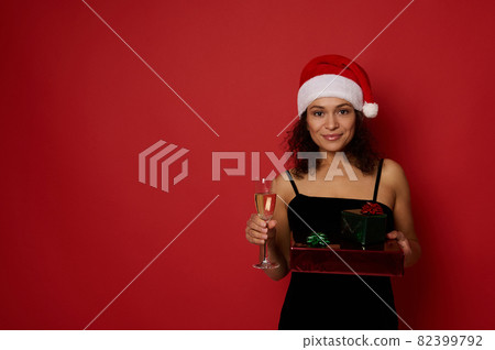 Beautiful Hispanic woman holds Christmas gift boxes wrapped in glitter red green gift paper and champagne flute with sparkling wine, poses on red background, cute smiling looking at camera. Copy space 82399792