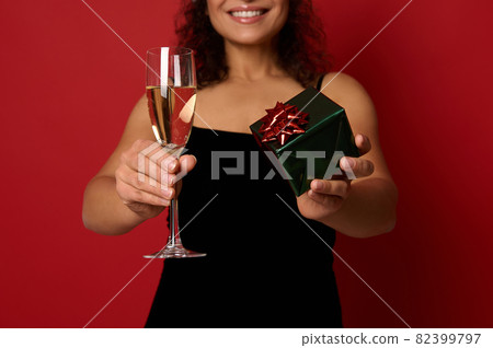 Soft focus on champagne flute with sparkling wine and box wrapped in glitter gift paper in the hands of pretty woman with beautiful toothy smile. Christmas concepts on red background with copy space Soft focus on champagne flute with sparkling wine and box wrapped in glitter gift paper in the hands of pretty woman with beautiful toothy smile. Christmas concepts on red background with copy space 82399797
