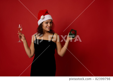 Pretty woman wearing Santa hat and black velvet dress holds champagne flute with sparkling wine and gift box, smiles toothy smile posing against red background, copy space. Christmas, Happy New Year Pretty woman wearing Santa hat and black velvet dress holds champagne flute with sparkling wine and gift box, smiles toothy smile posing against red background, copy space. Christmas, Happy New Year 82399798