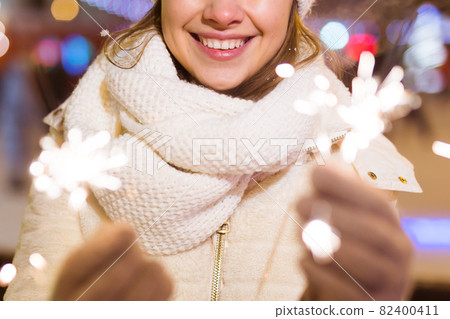 Young beautiful woman in knitted hat and scarf standing in the city with bengal light, sparkler. Concept celebration and christmas. Young beautiful woman in knitted hat and scarf standing in the city with bengal light, sparkler. Concept celebration and christmas. 82400411