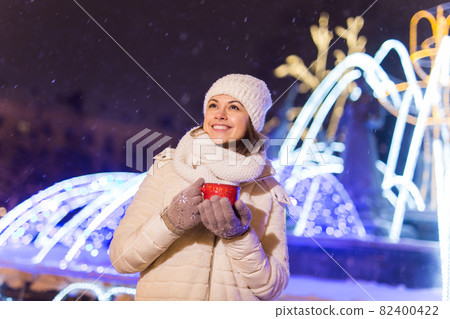 Christmas holidays. Beautiful smiling woman in warm clothing with cup of hot coffee outdoor in winter. City night. 82400422