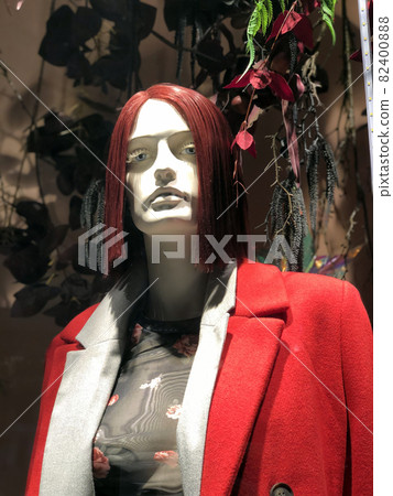 Woman's mannequin on the window of designer clothing store in Cyprus. Mannequin is wearing red jacket and translucent top. On the head is a burgundy wig with a bob hairstyle. Plants in the background Woman's mannequin on the window of designer clothing store in Cyprus. Mannequin is wearing red jacket and translucent top. On the head is a burgundy wig with a bob hairstyle. Plants in the background 82400888