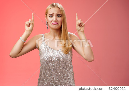 Upset whining immature spoiled blond rich girl in silver glittering dress pouting frowning gonna cry pointing up regret jealousy begging buy expensive shoes, standing red background displeased 82400911