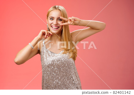 Charismatic funny amused charming blond girl 25s having fun dancing red background in silver trendy glittering party dress show peace disco gesture near eye smiling laughing carefree 82401142