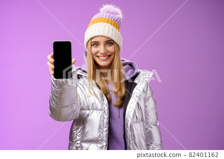 Friendly cheerful confident blond girl in silver stylish winter jacket hat extend arm showing smartphone display advertising awesome new device app smiling self-assured recommend use mobile phone 82401162