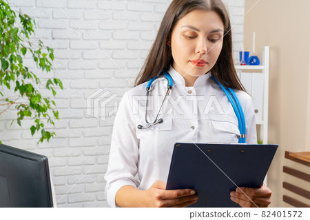 Young brunette woman doctor standing with clipboard in her office 82401572